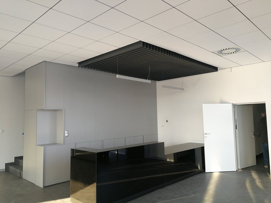 Nowoczesne biuro z czarnym błyszczącym blatem recepcyjnym, szara zabudowa meblowa, białe ściany i sufit kasetonowy z czarną podwieszaną konstrukcją nad recepcją.