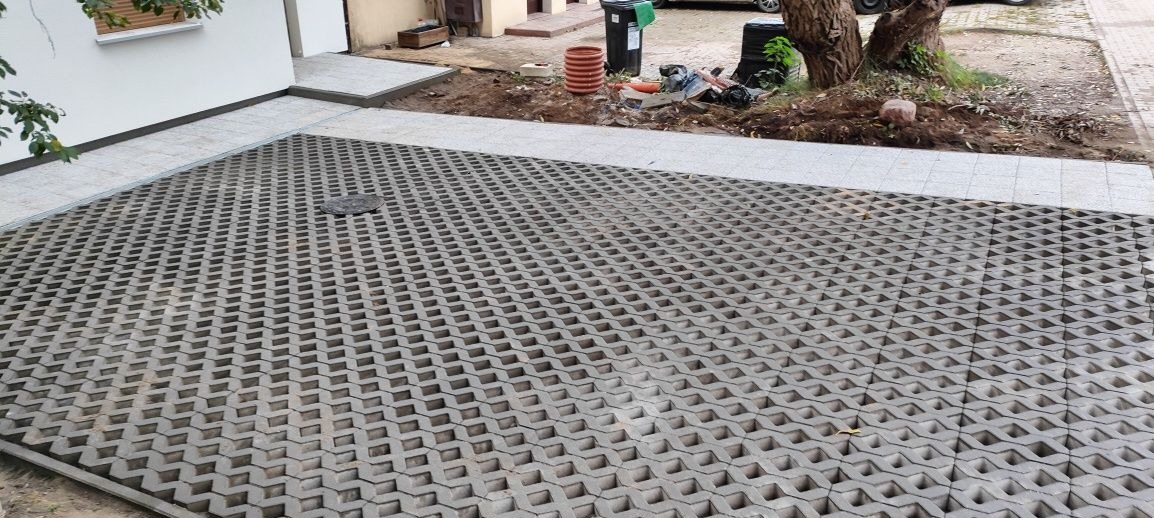 Ułożona szara ażurowa kostka brukowa na podjeździe w Poznaniu, obrzeża z jasnoszarych płyt, widoczny właz kanalizacyjny, w tle fragment posesji z zielenią.