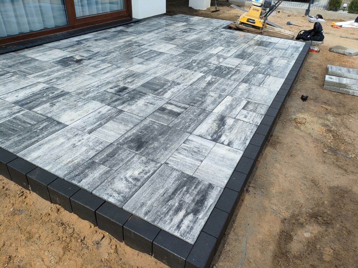 Ułożona szara kostka brukowa z czarnym obrzeżem na przygotowanym podłożu, widoczna zagęszczarka w tle, częściowo zakończona realizacja.