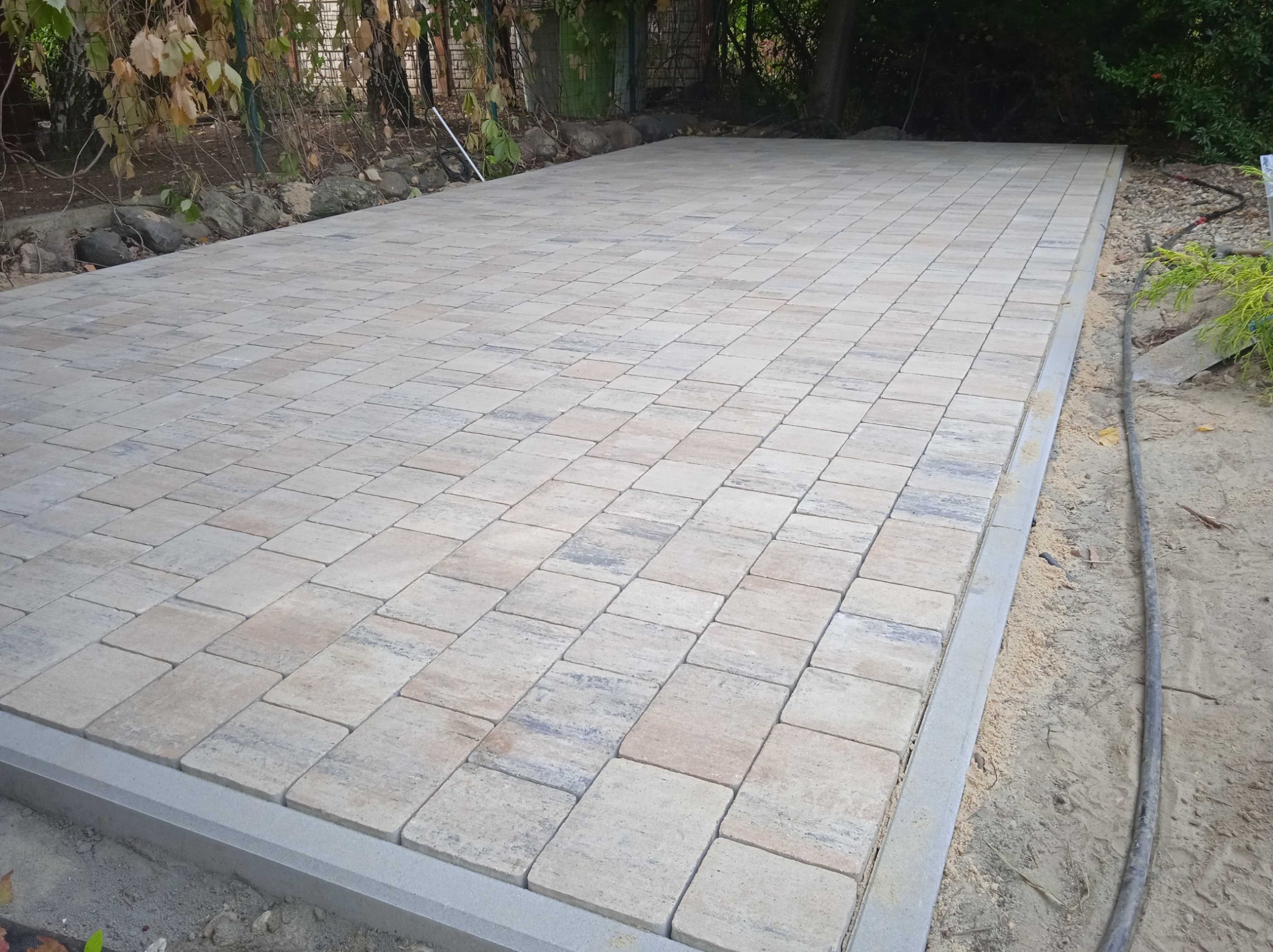 Ułożona nawierzchnia z kostki brukowej w odcieniach beżu i szarości, obrzeża wykończone betonową ramą, widoczne drzewa w tle.