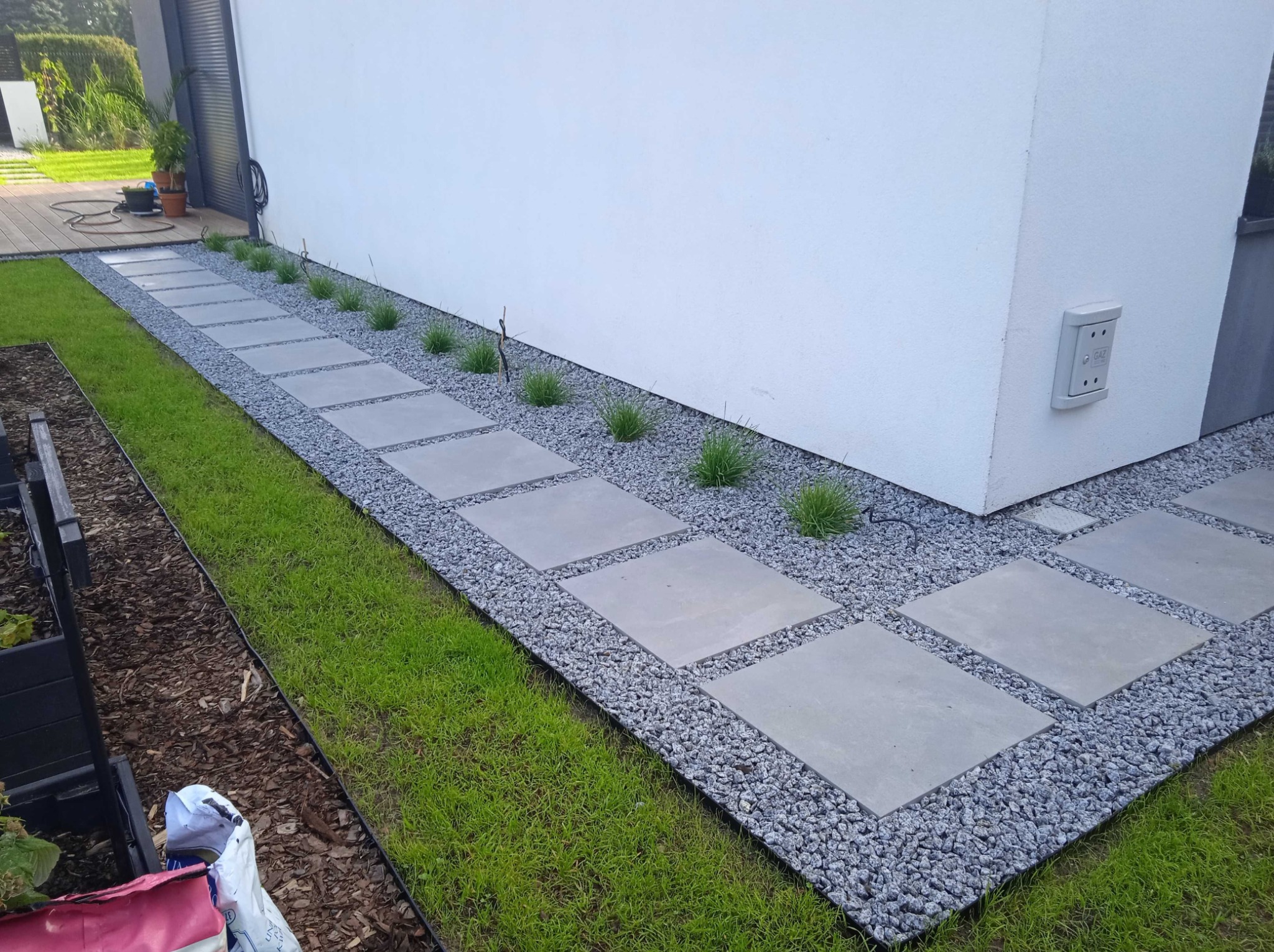 Ścieżka z kwadratowych płyt betonowych ułożona wzdłuż białej ściany budynku, otoczona szarym żwirem i zieloną trawą, z dodatkowymi nasadzeniami roślin.