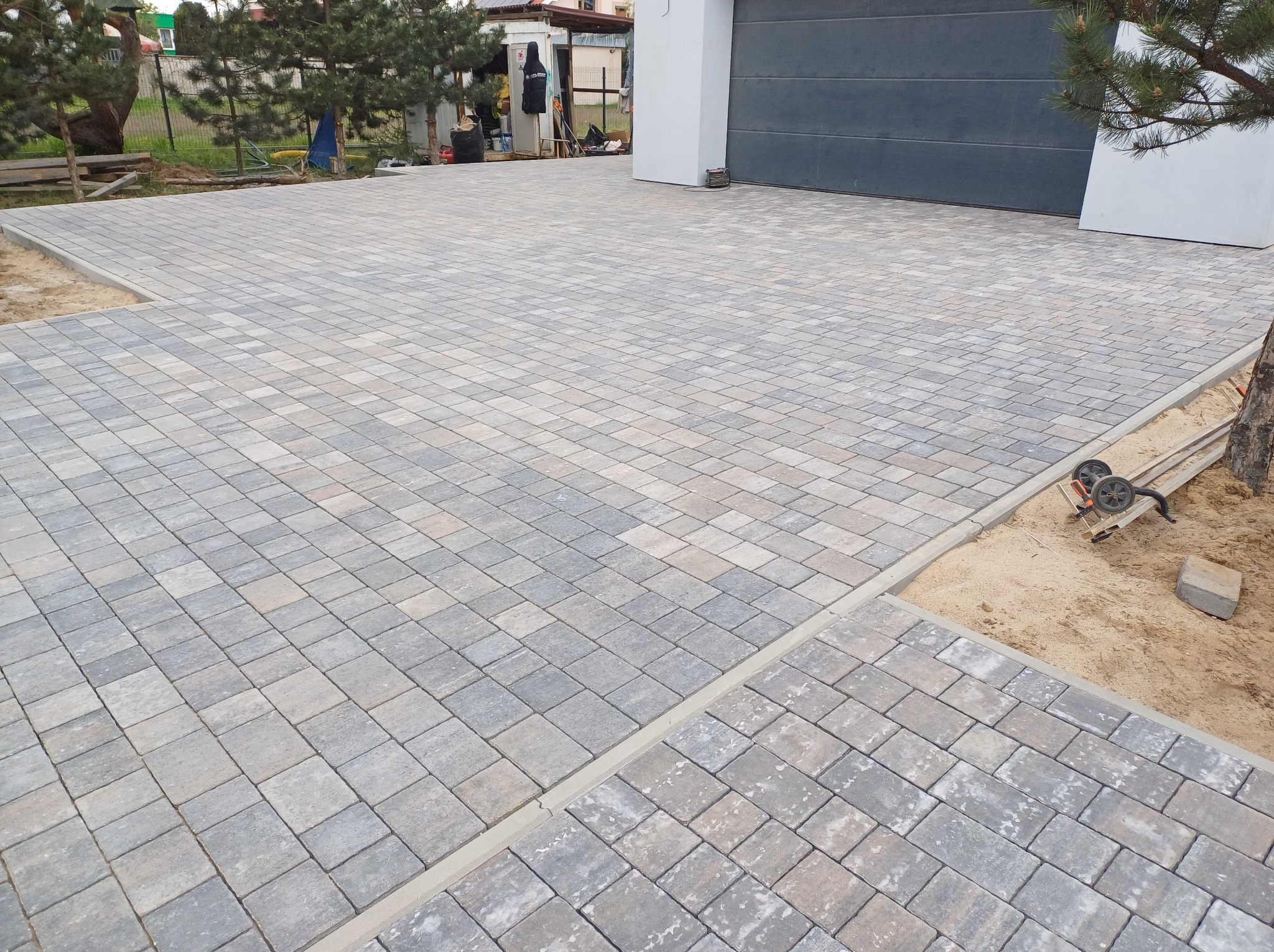 Ułożona, szara kostka brukowa na podjeździe przed garażem, widoczne obrzeża i piasek zasypujący szczeliny, ubijarka w tle.
