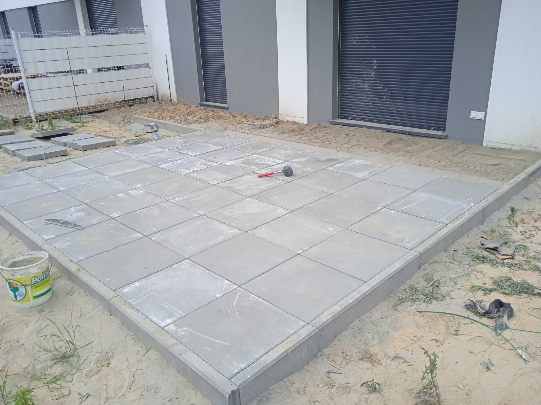 Niedokończona powierzchnia z szarej kostki brukowej ułożonej na piasku, obramowana szarym obrzeżem, widoczny gumowy młotek i poziomica, w tle budynek z szarymi roletami.