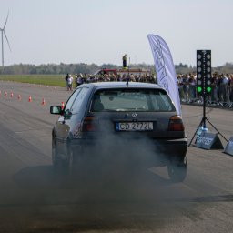 Uslugi fotograficzne - Czarny Volkswagen Golf z numerem rejestracyjnym GD 2772L pali gumę na starcie wyścigu drag racing, tło stanowi tłum widzów, sygnalizacja świetlna i wiatrak.