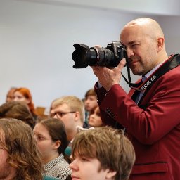 Uslugi fotograficzne - Mężczyzna w bordowej marynarce celuje aparatem Canon EOS 6D w stronę siedzącego tłumu, prawdopodobnie podczas konferencji lub wydarzenia.