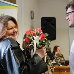 Uslugi fotograficzne - Uśmiechnięta kobieta w czarnej skórzanej kurtce trzyma bukiet róż, wręczając go uśmiechniętemu mężczyźnie w okularach i białej koszuli.