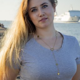 Uslugi fotograficzne - Portret kobiety z długimi blond włosami i naszyjnikiem w kształcie serc, na tle morza i statku pasażerskiego w oddali. Ujęcie z lekkim pochyleniem głowy.