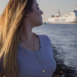 Uslugi fotograficzne - Profil kobiety z blond włosami i złotym naszyjnikiem w kształcie serca, na tle morza i statku Unity Line w oddali, uchwycony w miękkim świetle.