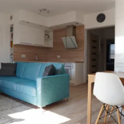 Jasne wnętrze mieszkania w Szczecinie: turkusowa sofa z poduszkami, drewniany stół z białymi krzesłami, kuchnia z drewnianymi akcentami i srebrnym okapem.