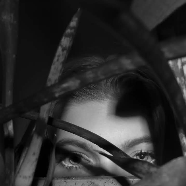 Portret kobiety w czerni i bieli, z widocznymi oczami spoglądającymi przez liście roślin doniczkowych, tworząc tajemniczy i intymny kadr.