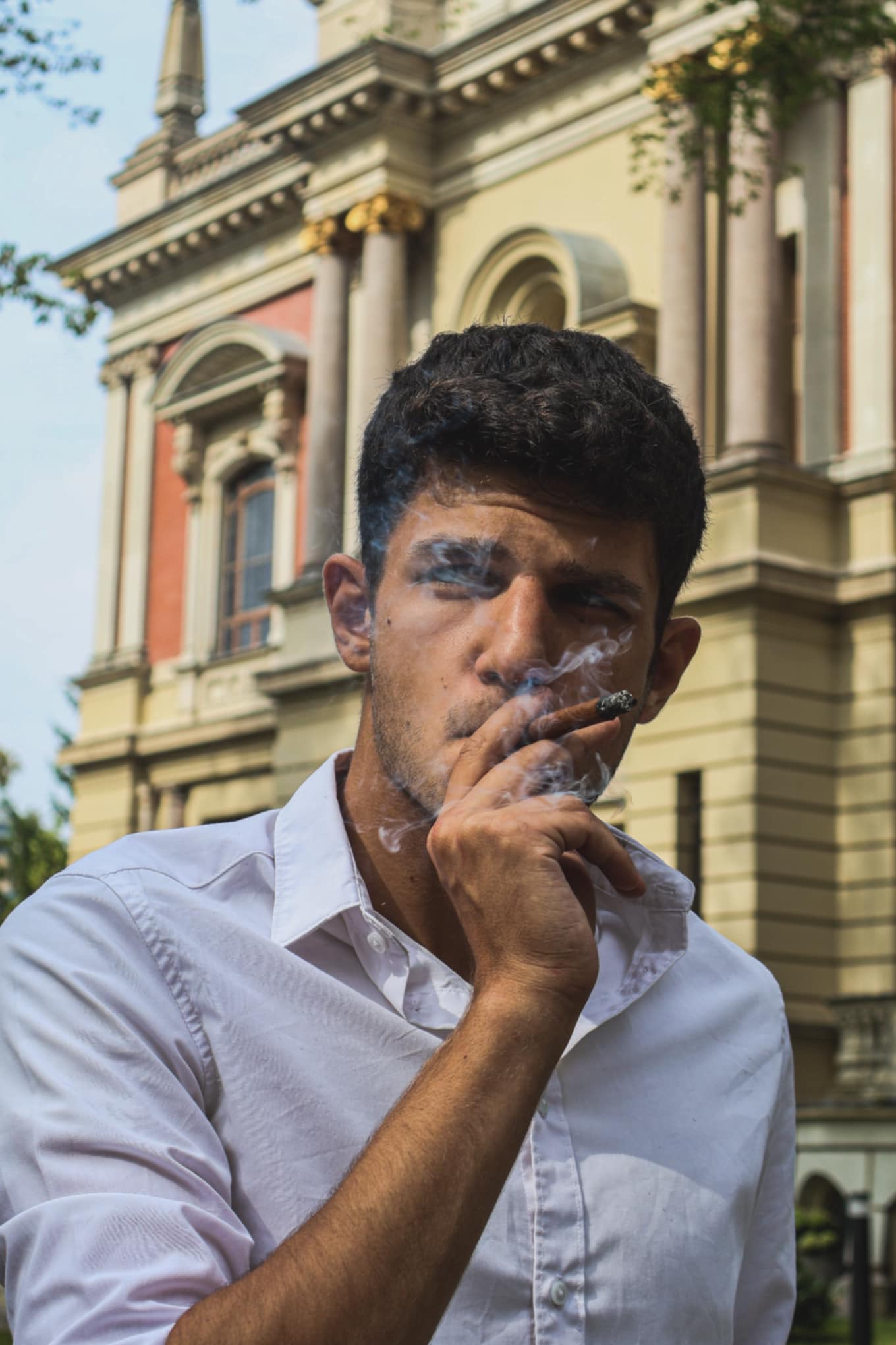 Portret młodego mężczyzny w białej koszuli, palącego cygaro na tle klasycystycznej architektury z kolumnami.
