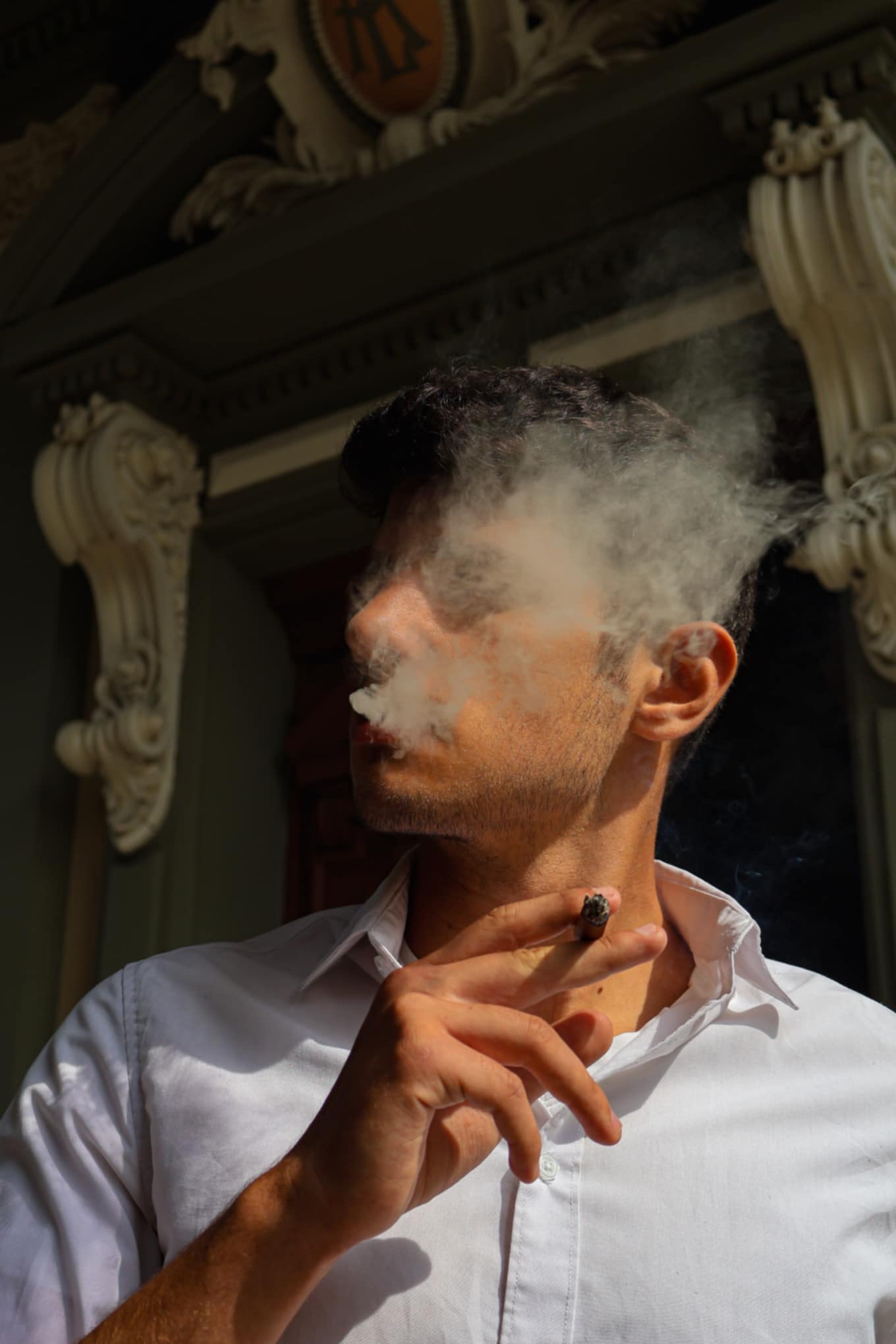 Mężczyzna w białej koszuli pali cygaro na tle zdobionej fasady budynku.