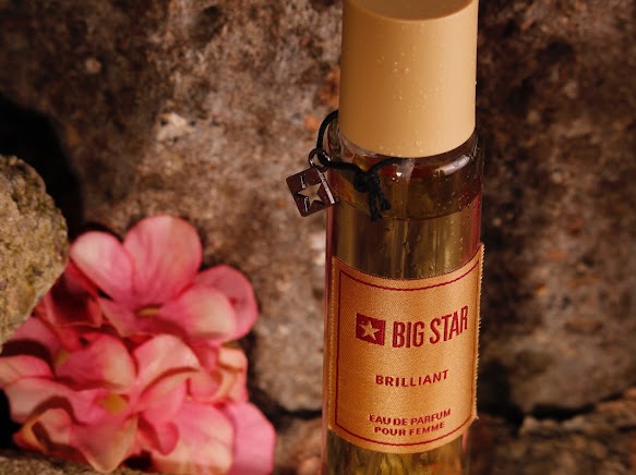 Butelka perfum Big Star Brillant na tle szarego kamienia i różowych kwiatów, zawieszka w kształcie gwiazdy przy szyjce.