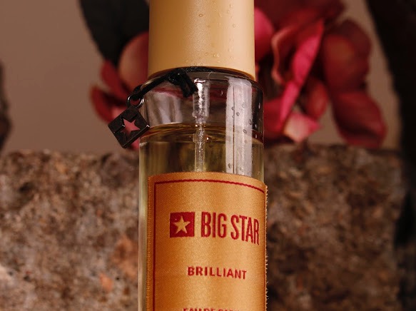 Butelka perfum Big Star Brillant z metalowym breloczkiem w kształcie gwiazdy, na tle kamienia i kwiatu.