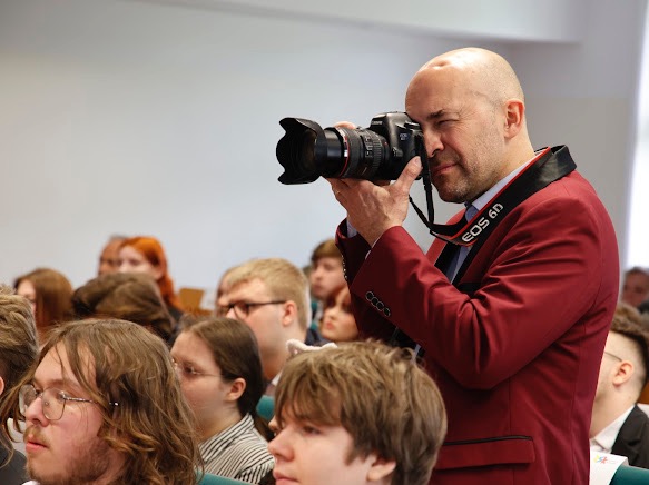 Mężczyzna w bordowej marynarce celuje aparatem Canon EOS 6D w stronę siedzącego tłumu, prawdopodobnie podczas konferencji lub wydarzenia.