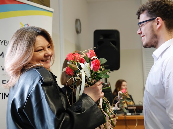 Uśmiechnięta kobieta w czarnej skórzanej kurtce trzyma bukiet róż, wręczając go uśmiechniętemu mężczyźnie w okularach i białej koszuli.