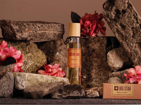 Flakon perfum Big Star Brilliant na tle kamieni i różowych kwiatów, z pudełkiem produktu w prawym dolnym rogu.