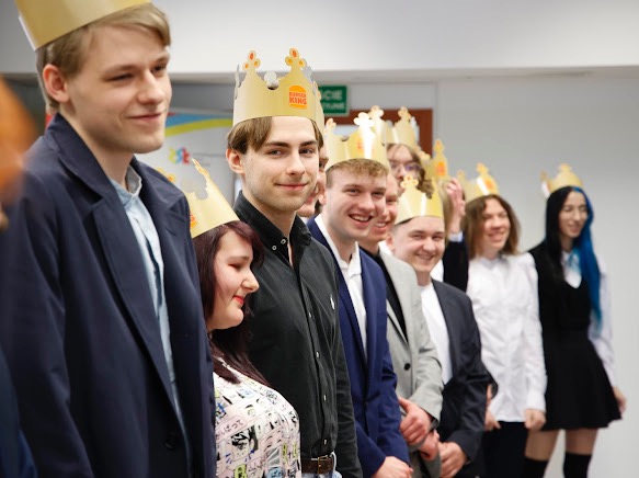 Grupa młodych ludzi w koronach Burger King ustawiona w rzędzie, uśmiechnięta, w formalnych strojach, prawdopodobnie podczas wydarzenia firmowego lub uroczystości.
