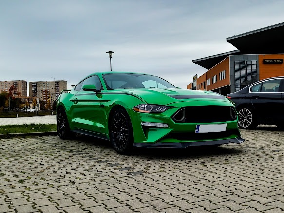 Zielony Ford Mustang z czarnymi akcentami zaparkowany na kostce brukowej w Szczecinie, obok stoi czarny samochód. W tle widoczny budynek.