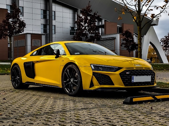 Żółte Audi R8 z czarnymi felgami na tle nowoczesnego budynku z elementami brązowego drewna i drzew z opadającymi liśćmi.