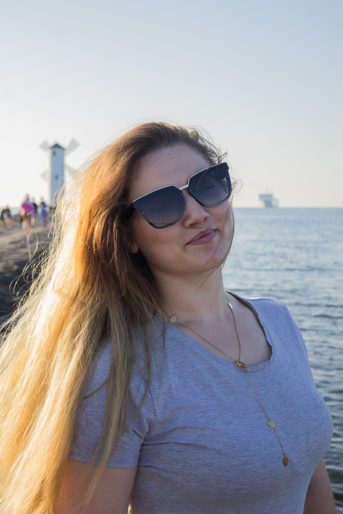 Portret kobiety w okularach na tle morza z wiatrakiem, jasne włosy, delikatny uśmiech, naszyjnik z serduszkami, naturalne światło, subtelny kadr.