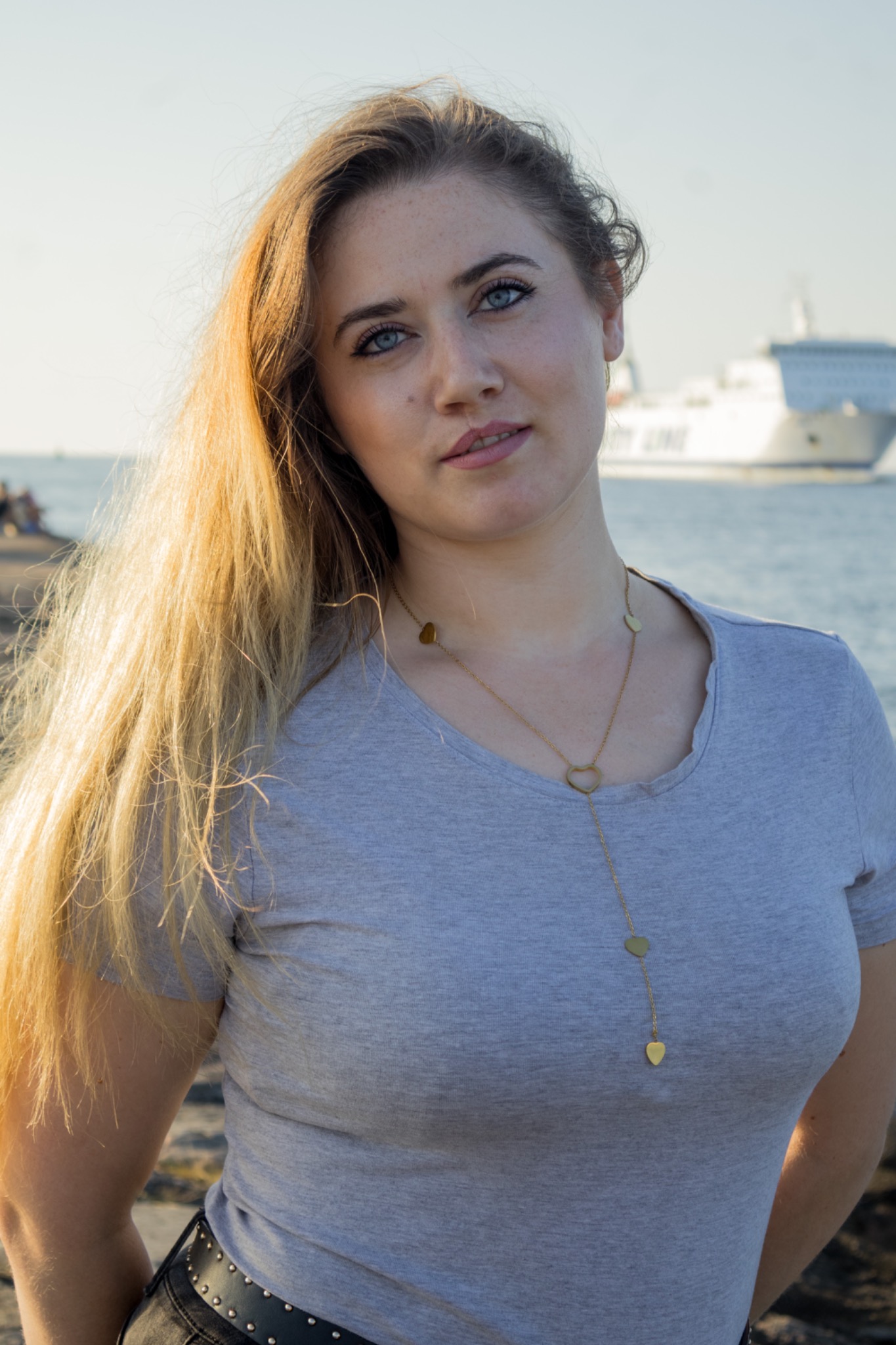 Portret kobiety z długimi blond włosami i naszyjnikiem w kształcie serc, na tle morza i statku pasażerskiego w oddali. Ujęcie z lekkim pochyleniem głowy.