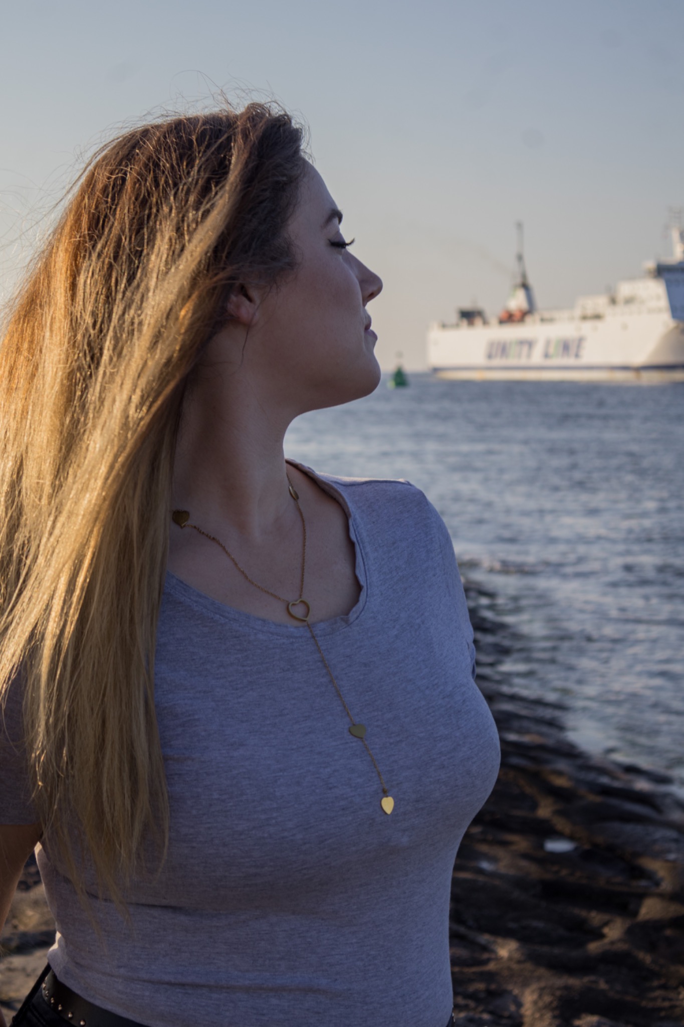 Profil kobiety z blond włosami i złotym naszyjnikiem w kształcie serca, na tle morza i statku Unity Line w oddali, uchwycony w miękkim świetle.