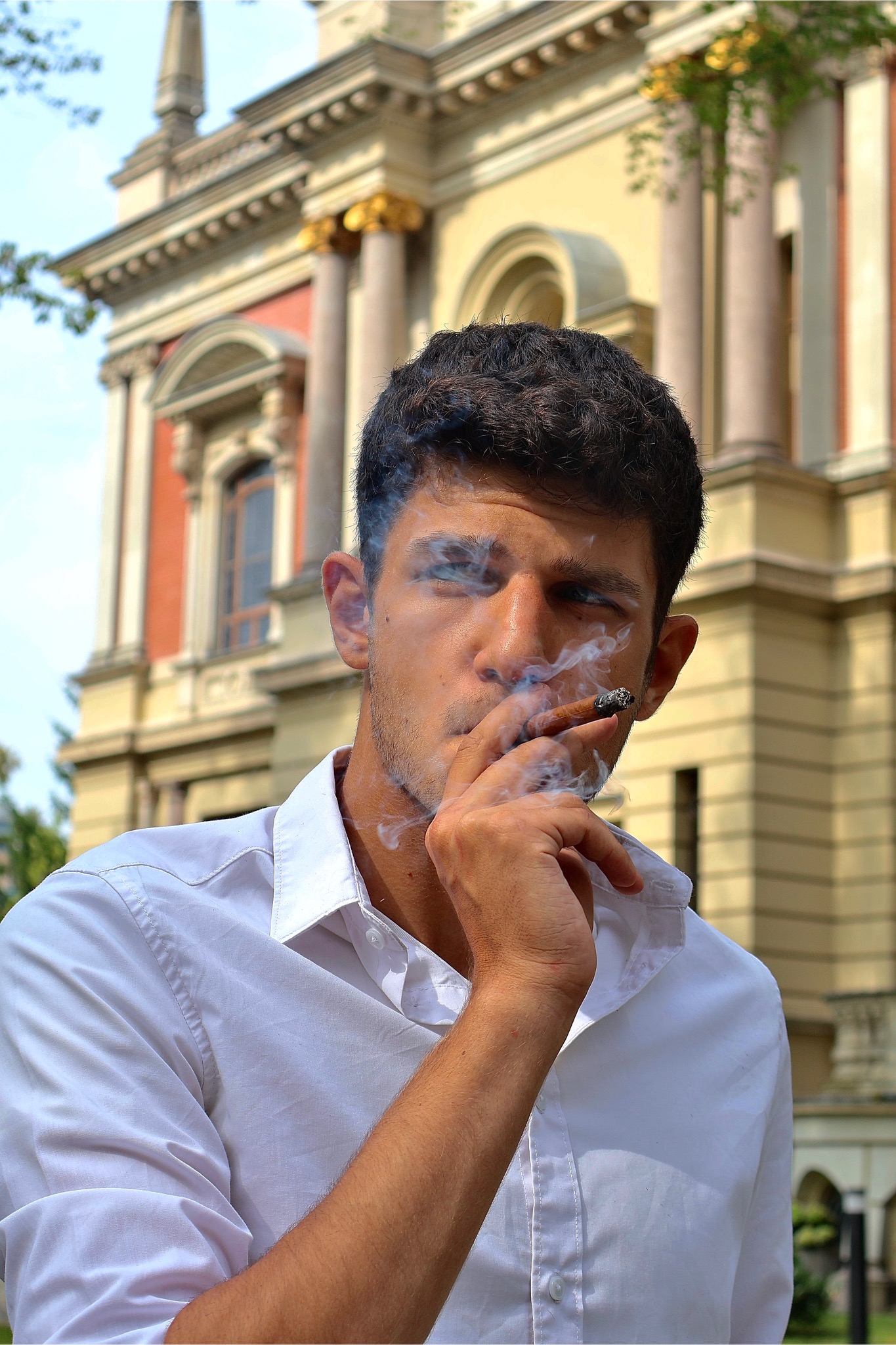 Mężczyzna w białej koszuli pali cygaro na tle klasycystycznego budynku z kolumnami. Dym otacza jego twarz, tworząc artystyczny efekt.