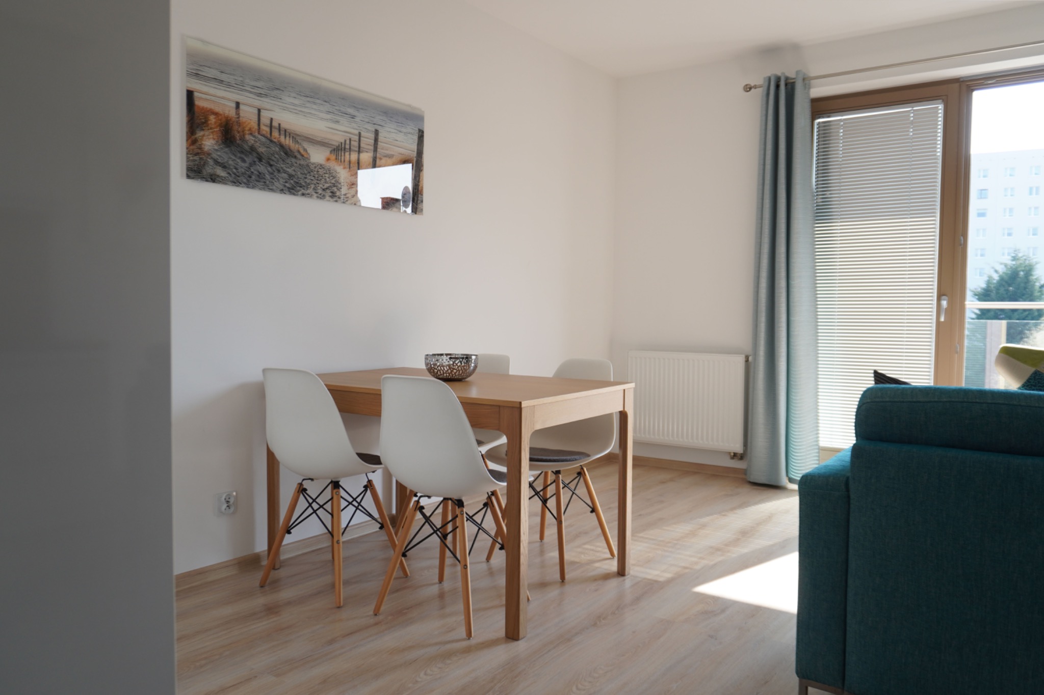 Jasne wnętrze z drewnianym stołem i białymi krzesłami. Na ścianie obraz z motywem plaży. Okno z żaluzjami i turkusowa zasłona. Minimalistyczny design.