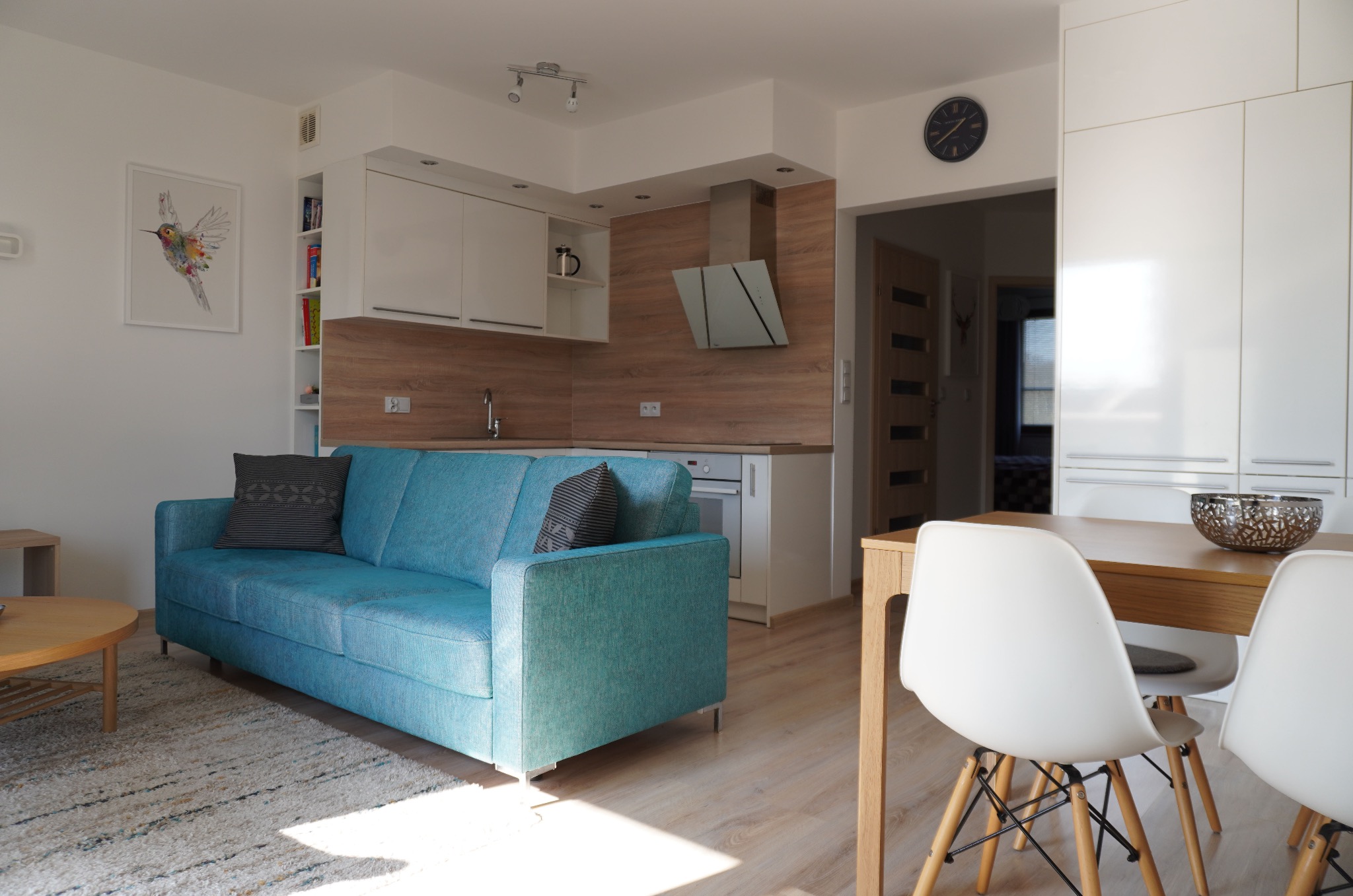Jasne wnętrze mieszkania w Szczecinie: turkusowa sofa z poduszkami, drewniany stół z białymi krzesłami, kuchnia z drewnianymi akcentami i srebrnym okapem.