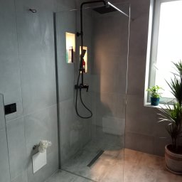 Flizer Rafał Jarek - Nowoczesna łazienka z szarą, płytkowaną ścianą, kabiną prysznicową typu walk-in z czarną armaturą, wnęką z oświetleniem i kosmetykami oraz roślinami doniczkowymi na parapecie okna.