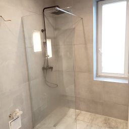 Flizer Rafał Jarek - Szklana kabina prysznicowa walk-in z czarną armaturą, szare płytki podłogowe i ścienne, okno z białymi ramami, widoczne niedokończone podłączenie elektryczne na ścianie.