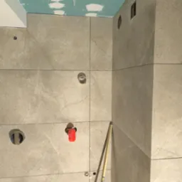 Ściana w trakcie układania płytek w łazience w Sosnowcu, widoczne otwory na instalacje, poziomica przy krawędzi i zielona linia lasera wyznaczająca poziom, sufit z płyt kartonowo-gipsowych...