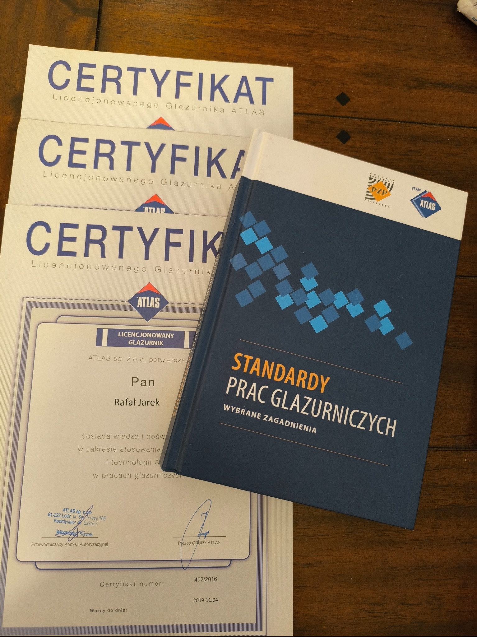 Stos certyfikatów licencjonowanego glazurnika Atlas oraz książka 'Standardy prac glazurniczych. Wybrane zagadnienia' na drewnianym stole.