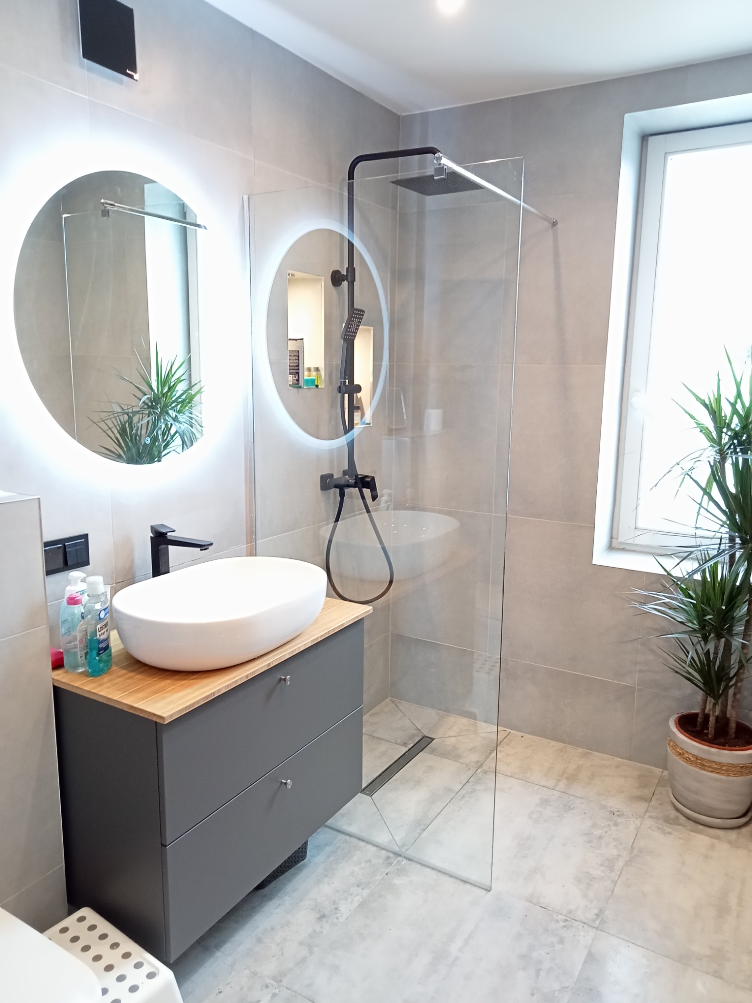 Nowoczesna łazienka z szarymi płytkami, owalną umywalką nablatową na szafce z drewnianym blatem, okrągłym lustrem z podświetleniem LED i kabiną prysznicową typu walk-in z czarną armaturą.
