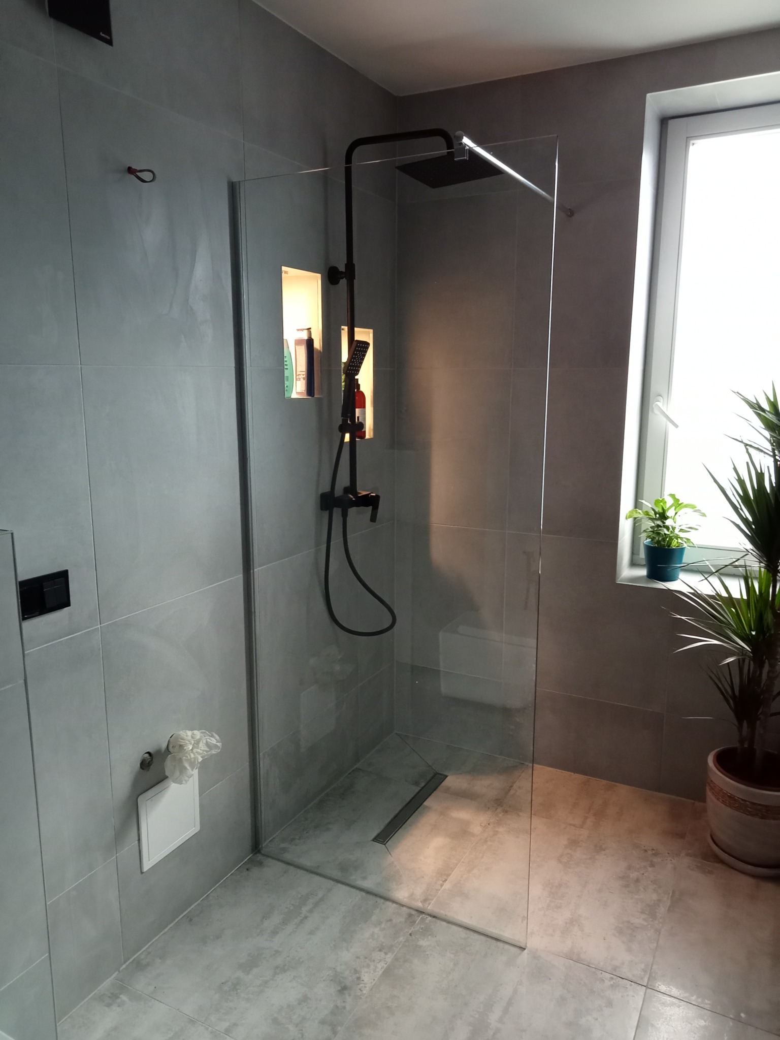 Nowoczesna łazienka z szarą, płytkowaną ścianą, kabiną prysznicową typu walk-in z czarną armaturą, wnęką z oświetleniem i kosmetykami oraz roślinami doniczkowymi na parapecie okna.