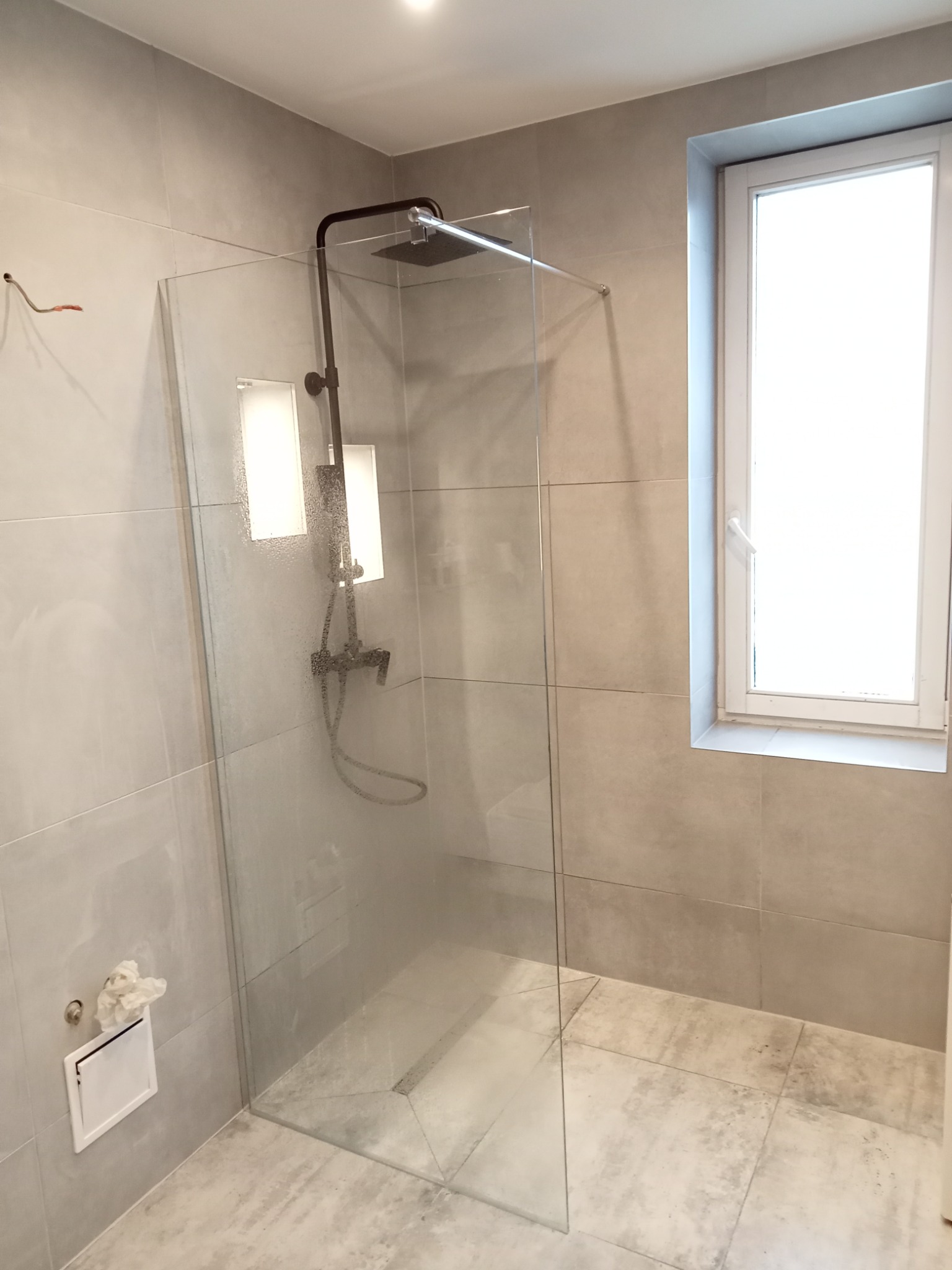 Szklana kabina prysznicowa walk-in z czarną armaturą, szare płytki podłogowe i ścienne, okno z białymi ramami, widoczne niedokończone podłączenie elektryczne na ścianie.