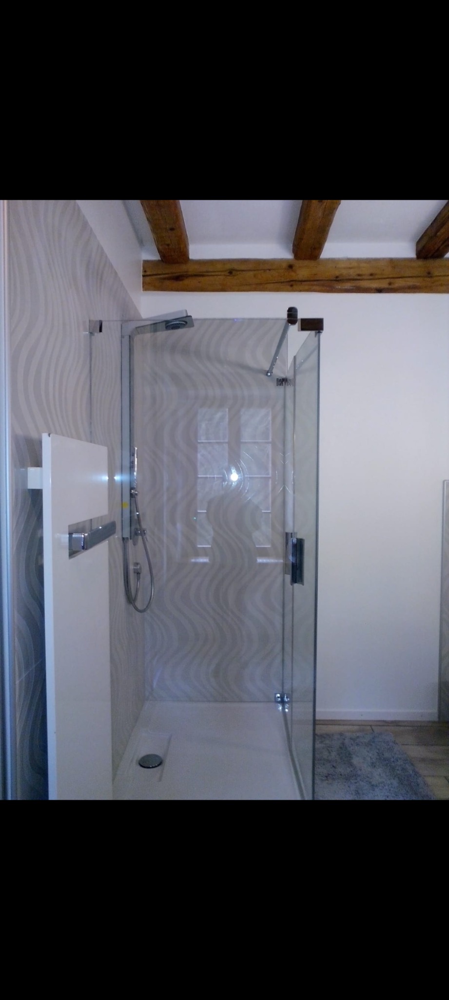 Nowoczesna kabina prysznicowa z deszczownicą i odpływem liniowym, ściany wykończone płytkami z motywem fal, drewniane belki stropowe.