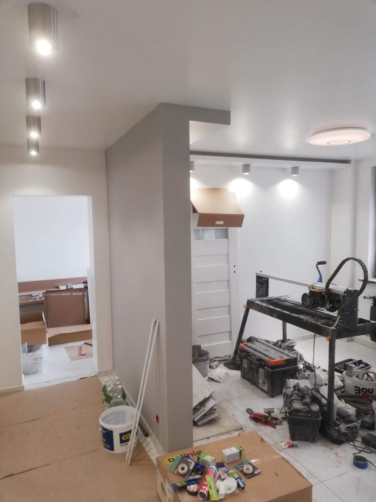 Wnętrze w trakcie prac wykończeniowych: widoczna przecinarka do płytek, narzędzia glazurnicze, karton z akcesoriami, świeżo zabudowana ściana działowa i oświetlenie sufitowe w dwóch pomieszczeniach.