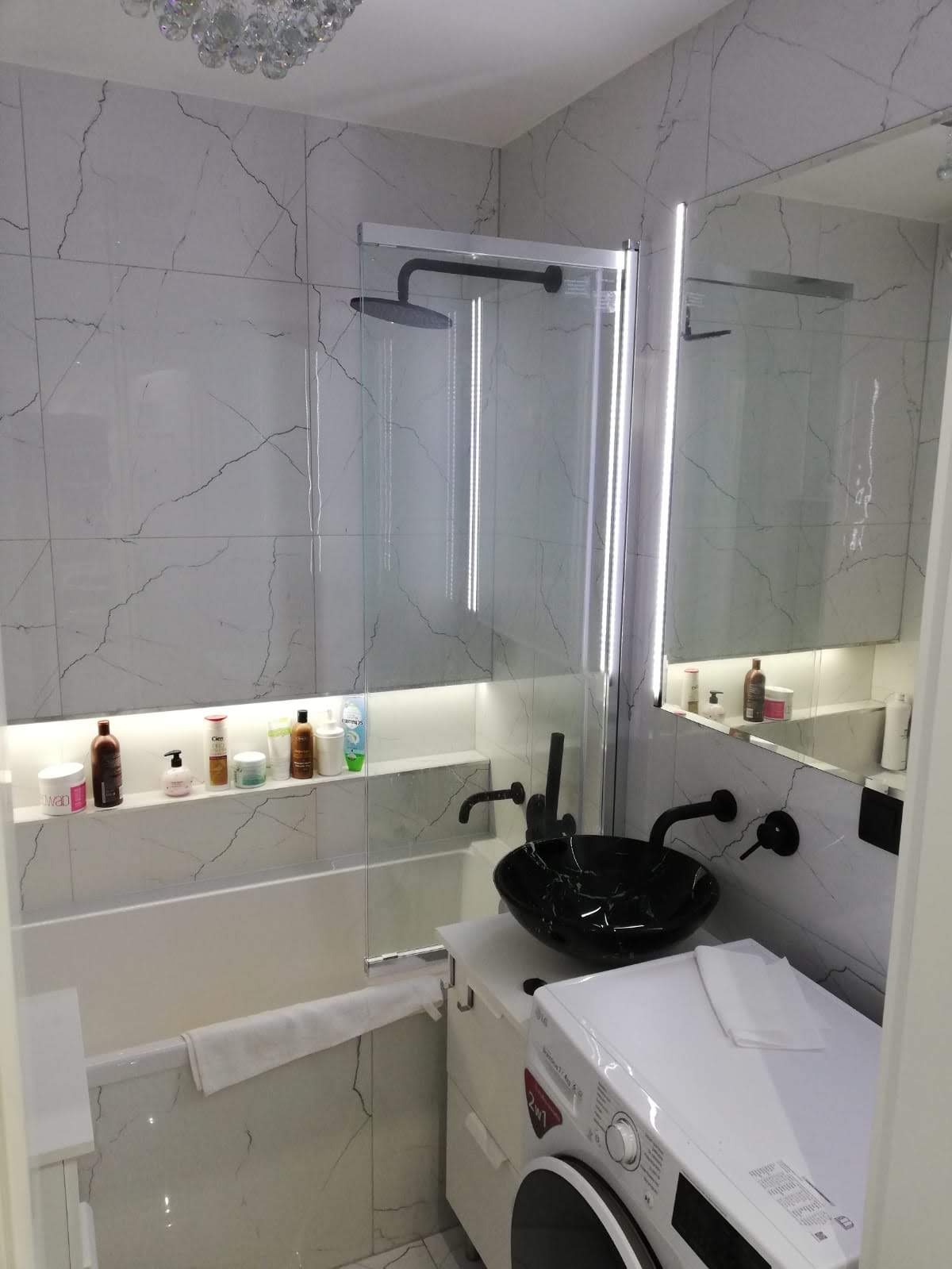 Nowoczesna łazienka z marmurowymi płytkami, czarną umywalką nablatową i oświetleniem LED w niszy nad wanną.