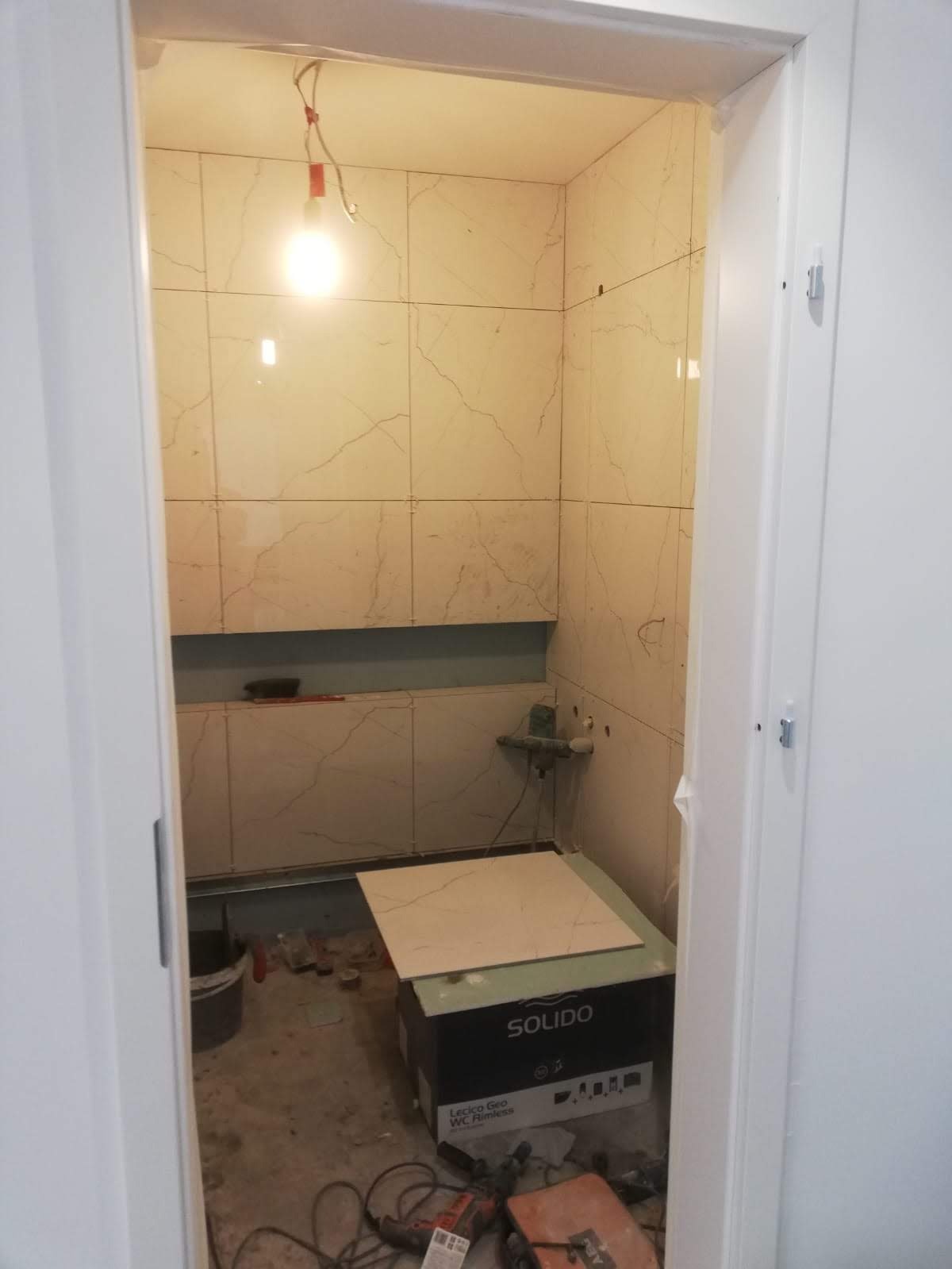 Remont łazienki w trakcie: ułożone marmurowe płytki ścienne, przygotowana pojedyncza płytka na pudełku po WC, widoczne narzędzia i prowizoryczne oświetlenie.