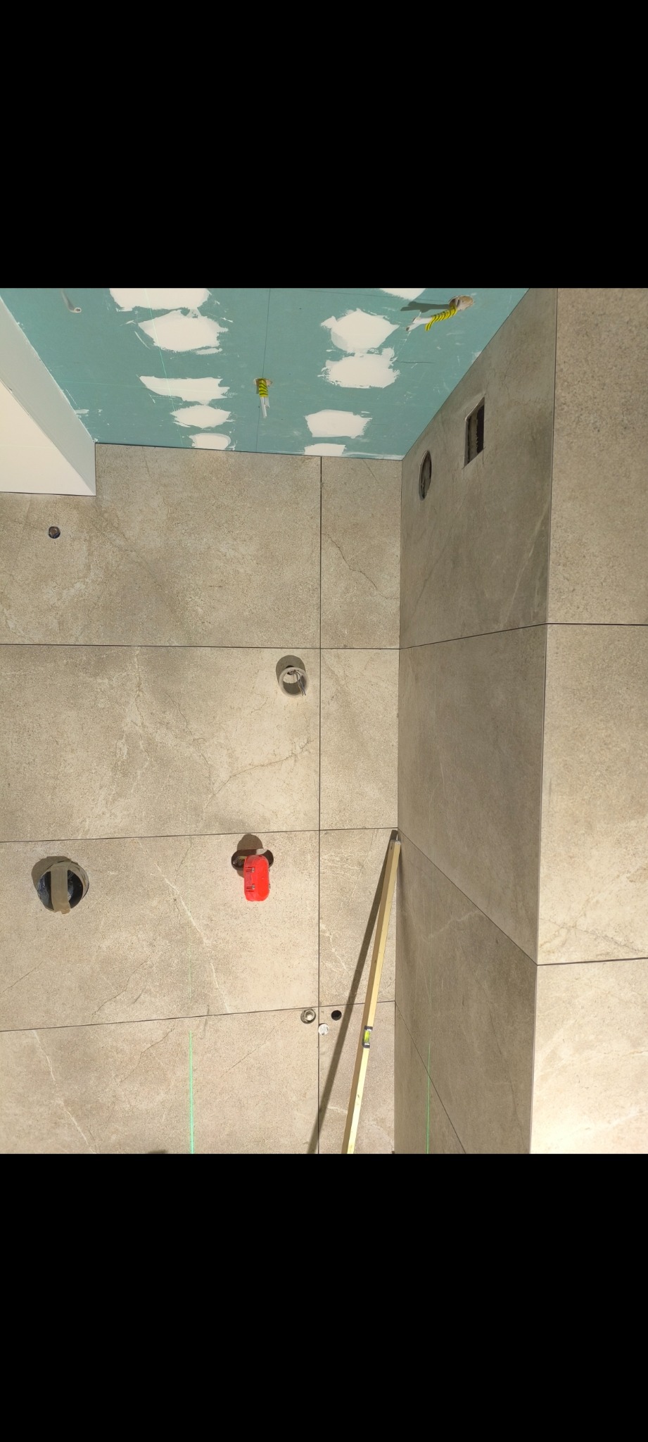 Ściana w trakcie układania płytek w łazience w Sosnowcu, widoczne otwory na instalacje, poziomica przy krawędzi i zielona linia lasera wyznaczająca poziom, sufit z płyt kartonowo-gipsowych...