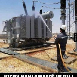 Elektro MP - Pracownik w kasku czyści transformator strumieniem wody z węża na zewnątrz, w tle słupy wysokiego napięcia i sucha trawa.