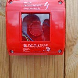 Elektro MP - Czerwony przycisk przeciwpożarowy wyłącznika prądu zamontowany na drewnianej powierzchni, widoczne oznaczenia i certyfikaty.