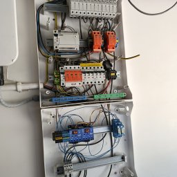 Elektro MP - Otwarta skrzynka elektryczna z zabezpieczeniami przepięciowymi, bezpiecznikami i przewodami. Widoczne elementy instalacji elektrycznej wewnątrz białej obudowy.