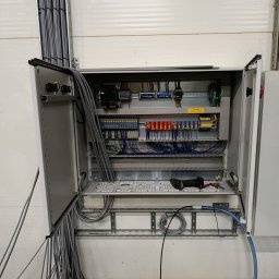 Elektro MP - Otwarta szafa sterownicza z widocznymi elementami instalacji elektrycznej, przewodami i narzędziami. Widoczne kable wchodzące do szafy.