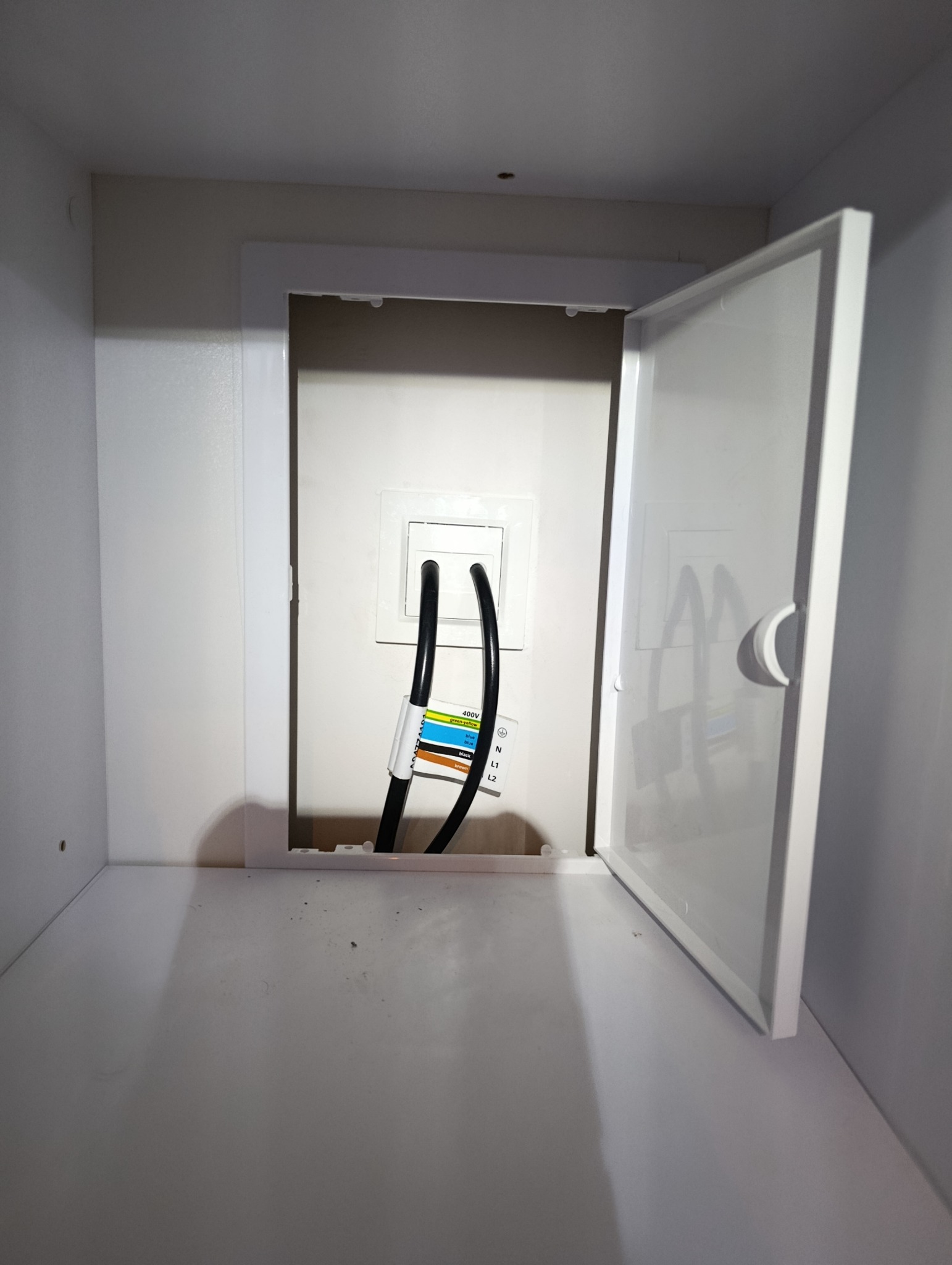 Biała szafka z otwartymi drzwiczkami, wewnątrz widoczne gniazdko elektryczne z podłączonymi przewodami. Widoczna etykieta z oznaczeniami kolorów przewodów.