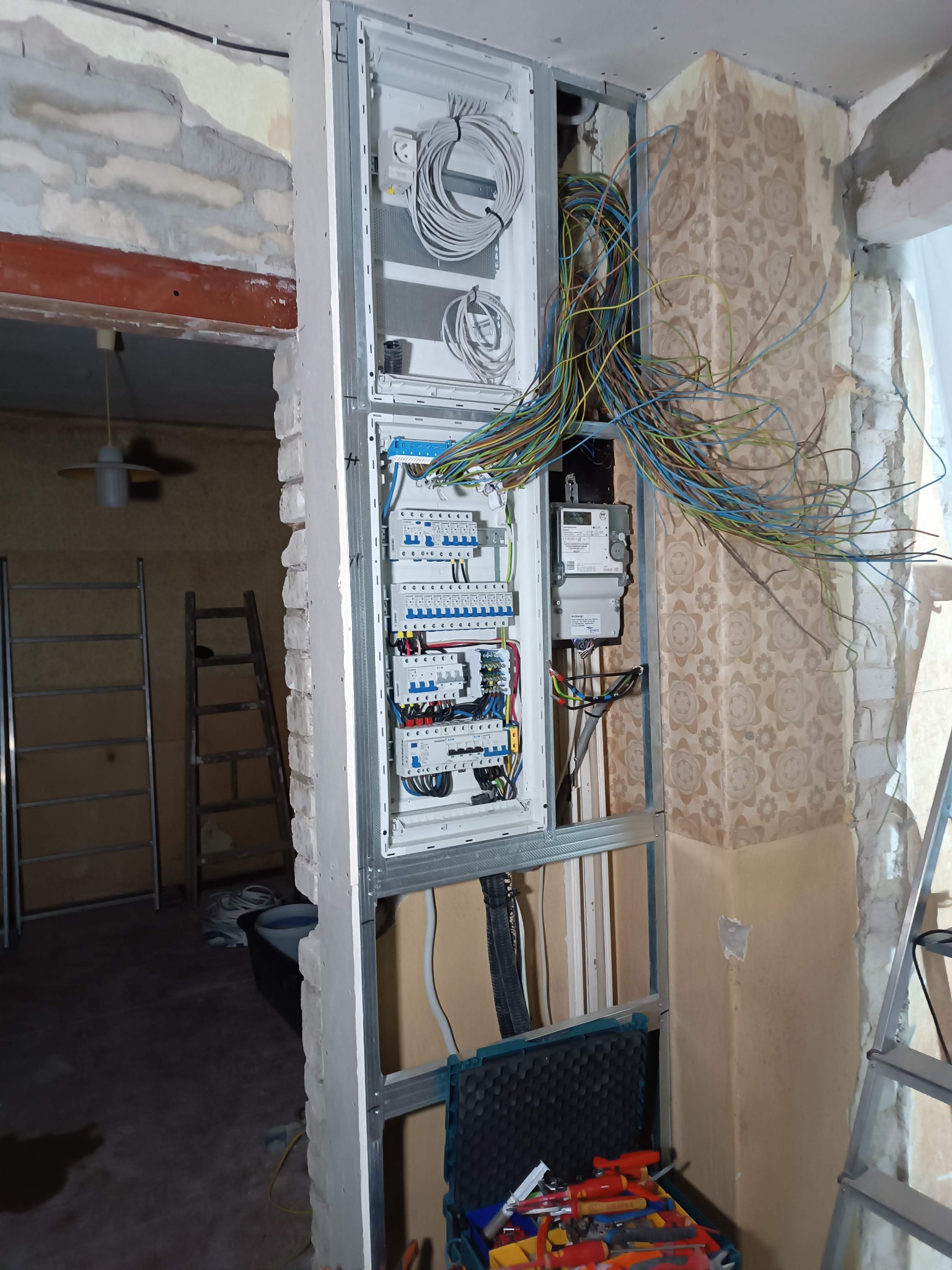 Wewnętrzna skrzynka elektryczna w trakcie montażu w remontowanym pomieszczeniu. Widoczne kable, liczniki i zabezpieczenia. W tle drabina i ściany z tapetą.