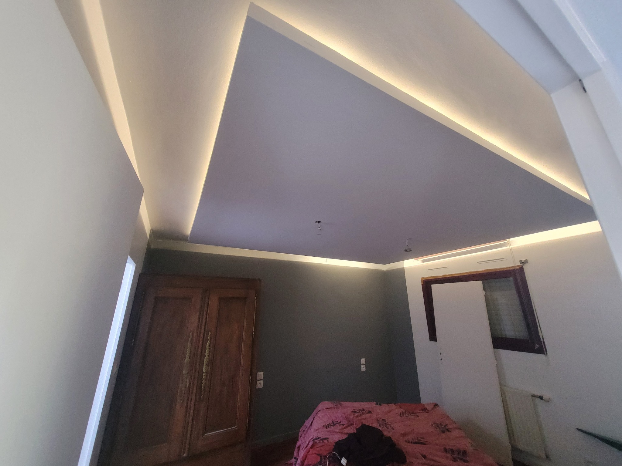 Pokój po odświeżeniu: szara ściana, drewniane drzwi, podświetlany sufit z wnękami, widoczne gniazdka elektryczne, łóżko z różową narzutą.