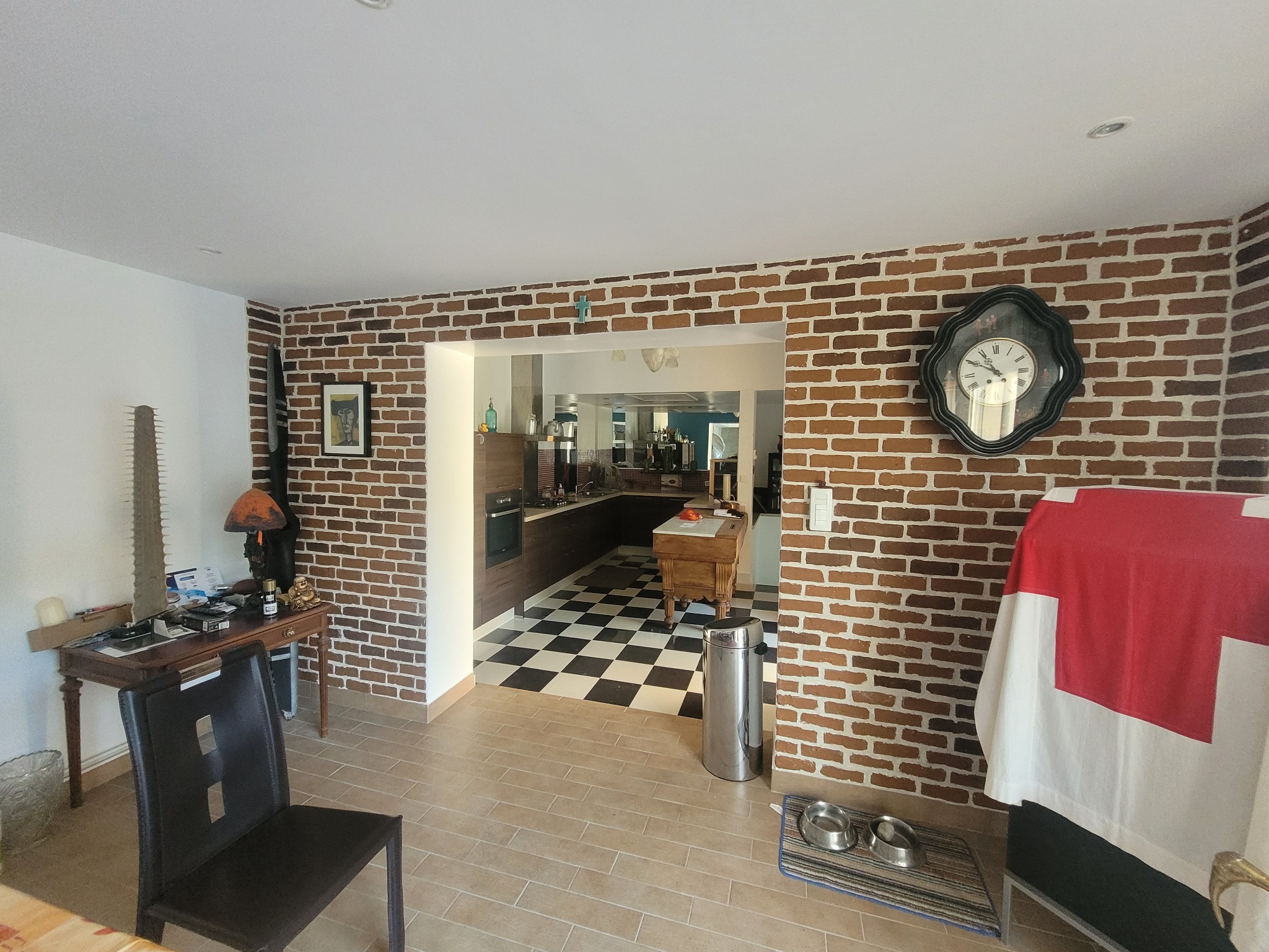 Wnętrze domu z ceglaną ścianą, widok na kuchnię z czarno-białą podłogą, na ścianie wiszący zegar w ozdobnej ramie, a na pierwszym planie krzesło i fragment stołu.